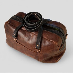Op de afbeelding: Een bruine leren reistas met zwarte leren handvatten en een bijpassende opgerolde zwarte schouderriem bovenop. De tas is voorzien van een ritssluiting aan de bovenzijde, koperkleurige metalen klinknagels en een groene voering.