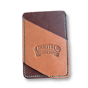 May include: A rectangular leather card holder featuring a dark brown base and a tan diagonal front pocket. The piece is stitched with teal thread and embossed with the text Corbett & Co. Leather Works in the center of the tan section.
