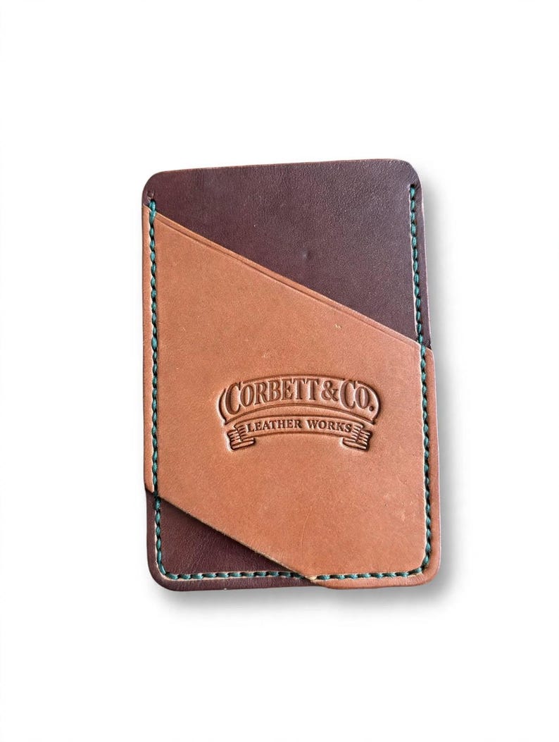 May include: A rectangular leather card holder featuring a dark brown base and a tan diagonal front pocket. The piece is stitched with teal thread and embossed with the text Corbett & Co. Leather Works in the center of the tan section.
