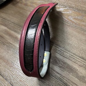 May include: A belt featuring a central black panel with a textured, quill-like pattern, bordered by smooth, deep magenta leather edges. The belt includes visible stitching along the borders and gold-toned metal hardware on the inner strap.