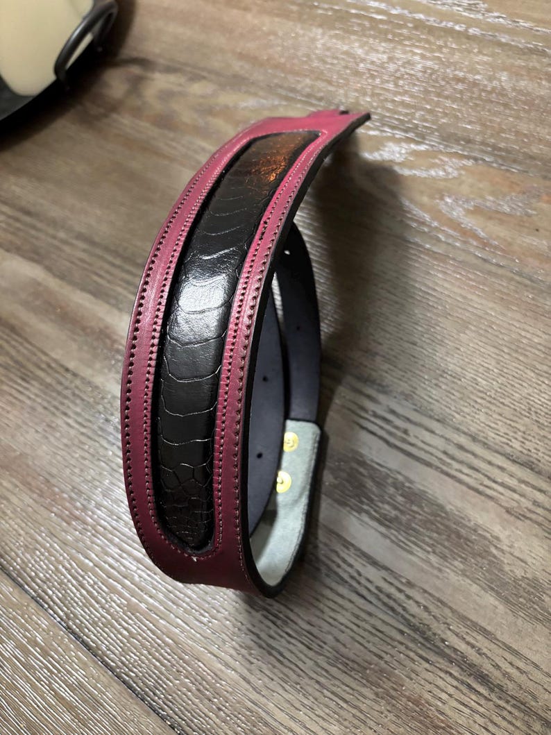 May include: A belt featuring a central black panel with a textured, quill-like pattern, bordered by smooth, deep magenta leather edges. The belt includes visible stitching along the borders and gold-toned metal hardware on the inner strap.
