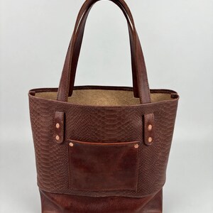 May include: A dark brown leather tote bag featuring a snakeskin-embossed texture on the upper body and a smooth leather base. The bag includes two sturdy handles, a front pocket, and copper-colored metal rivets securing the straps.