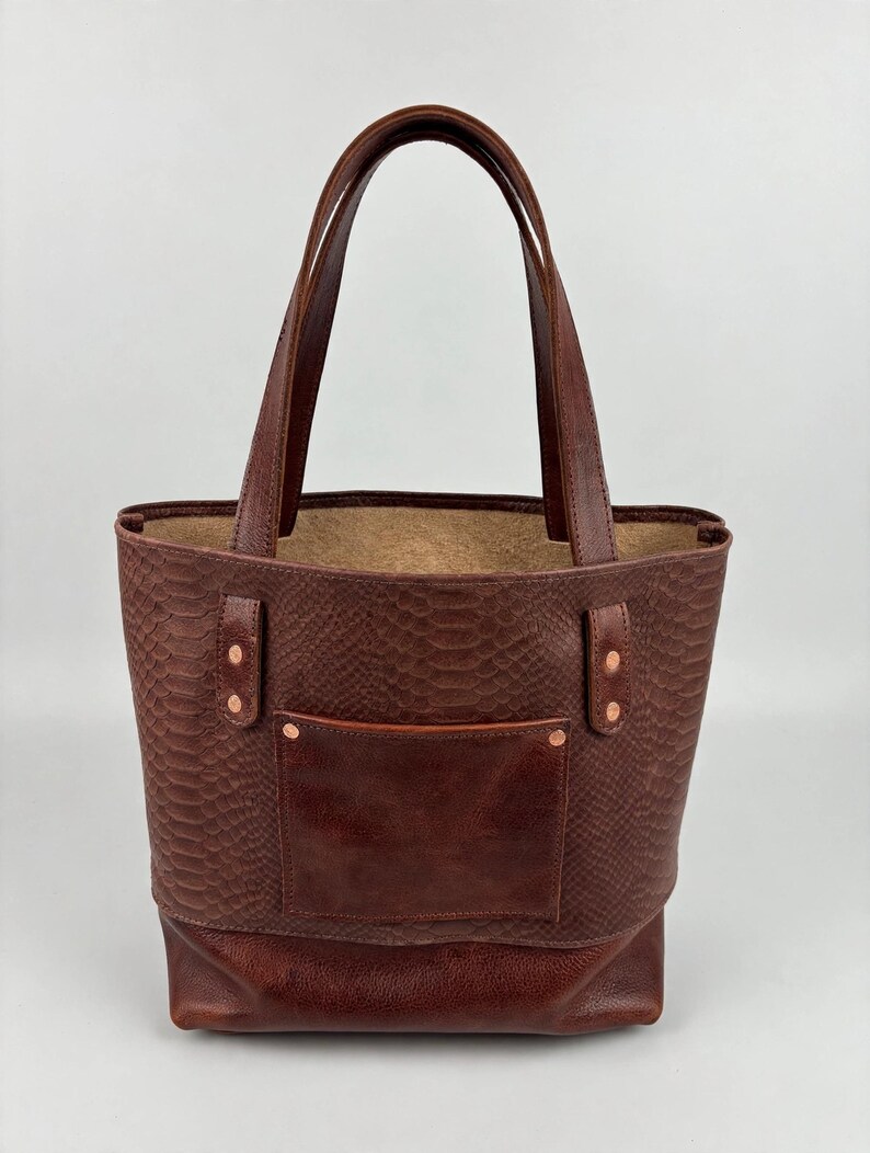 May include: A dark brown leather tote bag featuring a snakeskin-embossed texture on the upper body and a smooth leather base. The bag includes two sturdy handles, a front pocket, and copper-colored metal rivets securing the straps.