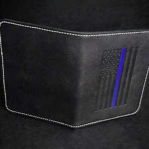Puede incluir: Una cartera de cuero negro tipo bifold con costuras blancas en contraste a lo largo de los bordes. La superficie frontal muestra un diseño grabado de la bandera estadounidense con una franja azul vertical en el centro.