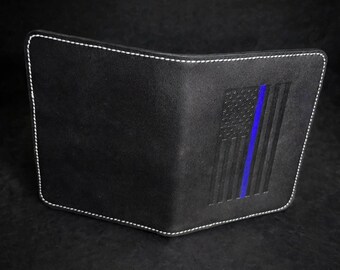 May include: A black leather bifold wallet featuring white contrast stitching along the edges. The front surface displays an embossed American flag design with a single vertical blue stripe running through the center of the flag.