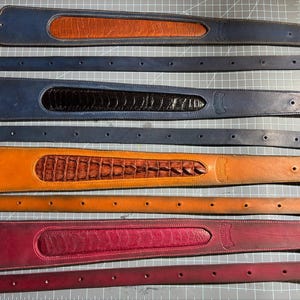 May include: Four sets of leather guitar straps, each paired with a matching thin leather belt. The straps feature wide, contoured ends with textured, exotic-style leather inlays in tan, black, brown, and magenta, with matching solid leather straps.