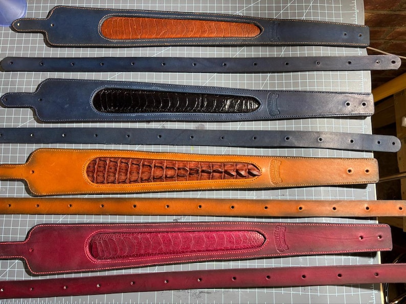 May include: Four sets of leather guitar straps, each paired with a matching thin leather belt. The straps feature wide, contoured ends with textured, exotic-style leather inlays in tan, black, brown, and magenta, with matching solid leather straps.