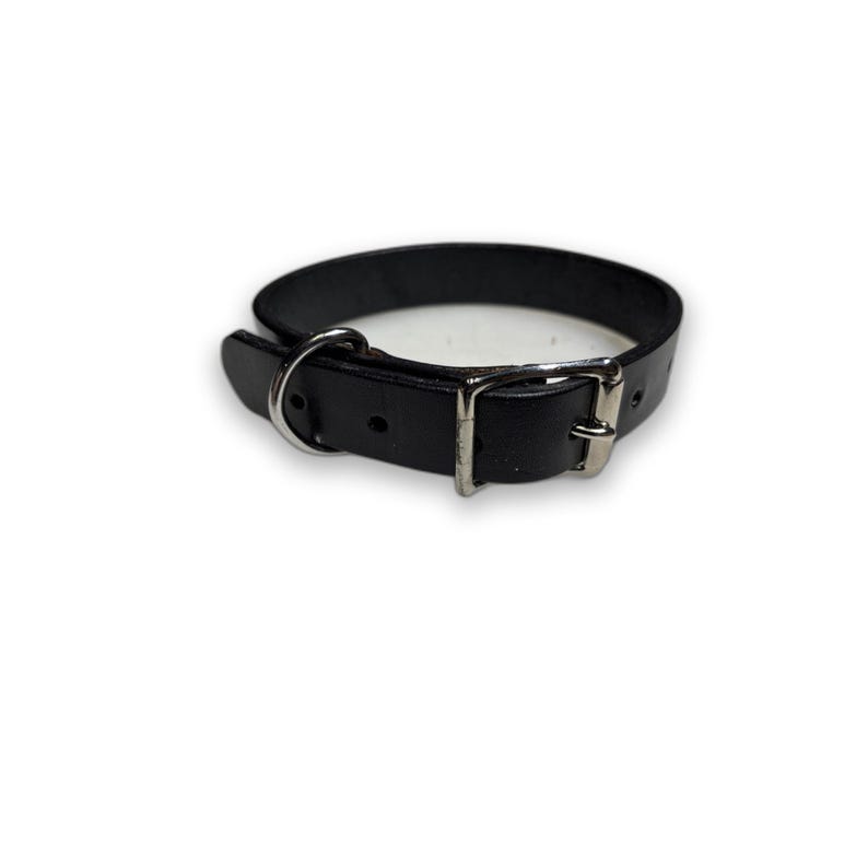 May include: A black leather collar featuring a silver-toned metal buckle, a rectangular loop, and a circular D-ring. The collar has multiple adjustment holes along the strap, providing a secure fit for the wearer with its classic design.