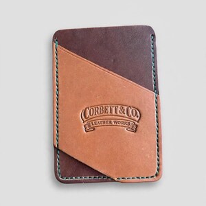 May include: A rectangular leather card holder featuring a dark brown base with a tan diagonal pocket overlay. The edges are stitched with teal thread. The tan section is embossed with the text Corbett & Co. Leather Works.