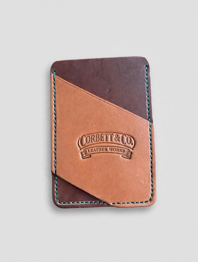May include: A rectangular leather card holder featuring a dark brown base with a tan diagonal pocket overlay. The edges are stitched with teal thread. The tan section is embossed with the text Corbett & Co. Leather Works.