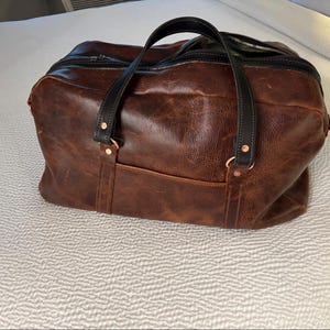 Op de afbeelding: Een bruine leren reistas met een getextureerd oppervlak, zwarte leren handvatten en een zwarte ritssluiting. De tas is voorzien van koperkleurige klinknagels die de handvatten aan de tas bevestigen en een voorvak aan de buitenzijde.