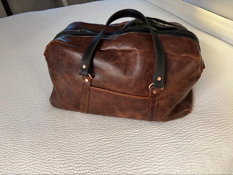Op de afbeelding: Een bruine leren reistas met een getextureerd oppervlak, zwarte leren handvatten en een zwarte ritssluiting. De tas is voorzien van koperkleurige klinknagels die de handvatten aan de tas bevestigen en een voorvak aan de buitenzijde.