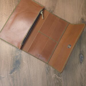Puede incluir: Una cartera larga de cuero color canela con costuras verdes en contraste, un compartimento con cremallera para monedas con tirador dorado, varias ranuras verticales para tarjetas y un bot&oacute;n a presi&oacute;n plateado.