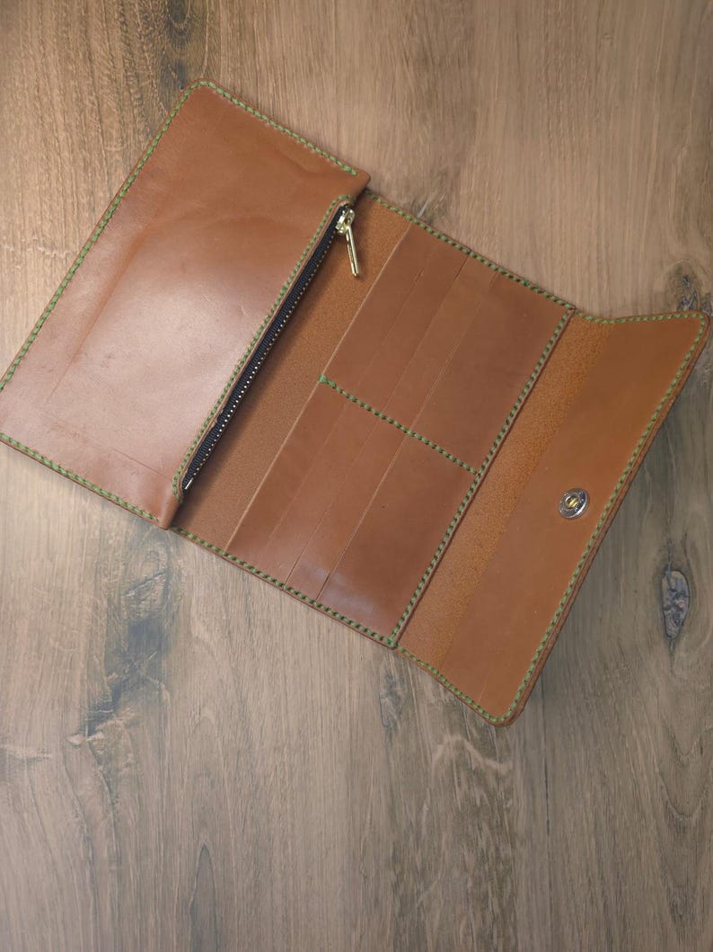 Puede incluir: Una cartera larga de cuero color canela con costuras verdes en contraste, un compartimento con cremallera para monedas con tirador dorado, varias ranuras verticales para tarjetas y un bot&oacute;n a presi&oacute;n plateado.