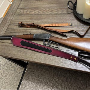 May include: A lever-action rifle with a wooden stock and metal barrel, accompanied by two rifle slings. One sling is made of plain tan leather with metal buckles, while the other is a wide, deep burgundy leather strap with a black center inlay.
