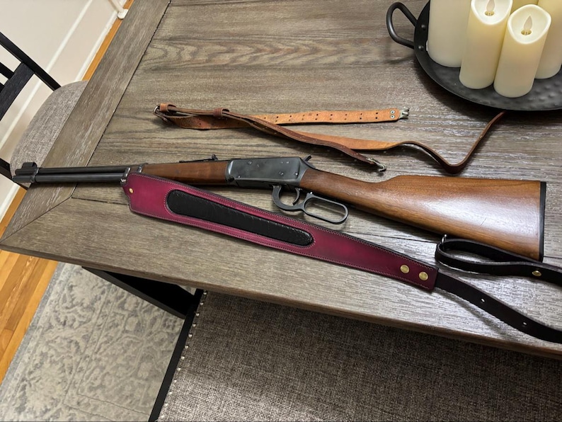 May include: A lever-action rifle with a wooden stock and metal barrel, accompanied by two rifle slings. One sling is made of plain tan leather with metal buckles, while the other is a wide, deep burgundy leather strap with a black center inlay.