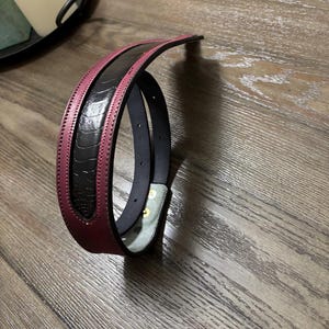 May include: A belt featuring a black textured center panel with a raised ostrich-like pattern, bordered by smooth deep burgundy leather edges. The belt includes visible stitching along the borders and two metallic snaps on the inner light-colored lining.