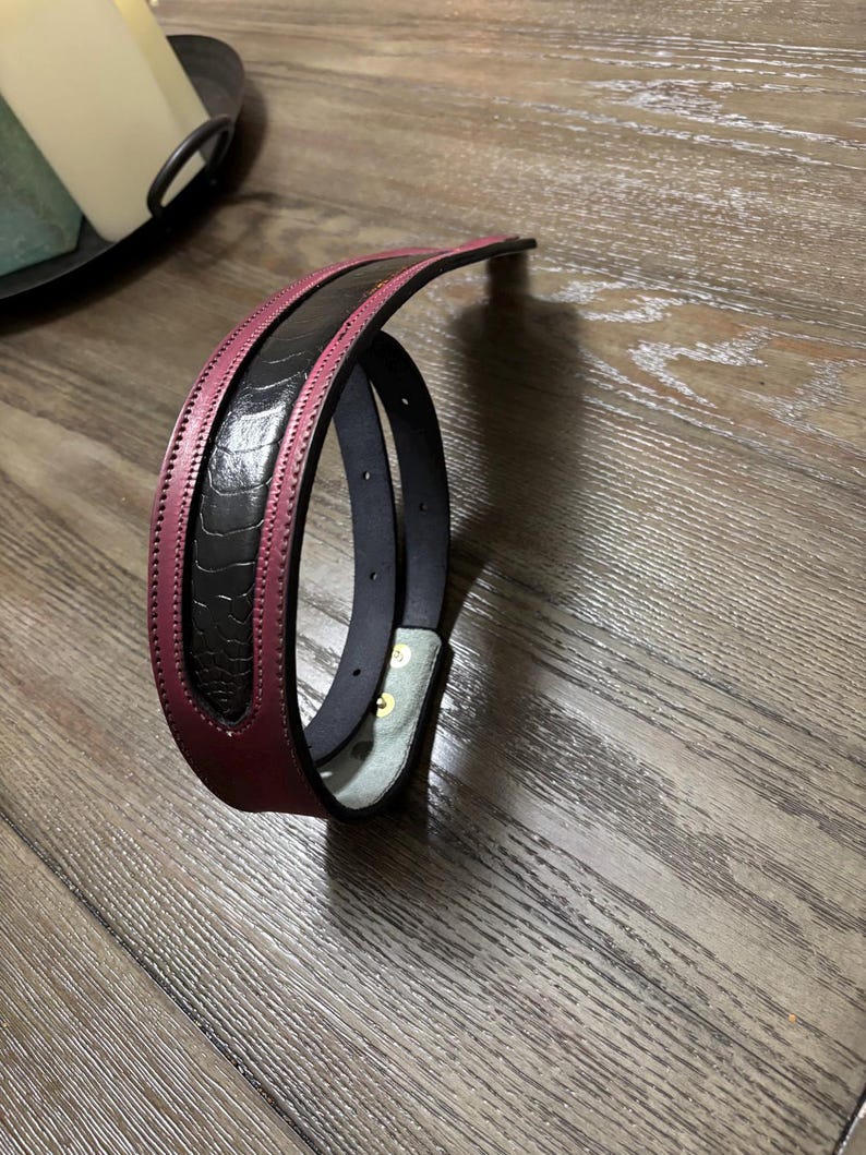 May include: A belt featuring a black textured center panel with a raised ostrich-like pattern, bordered by smooth deep burgundy leather edges. The belt includes visible stitching along the borders and two metallic snaps on the inner light-colored lining.