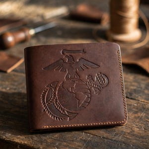 May include: A dark brown leather bifold wallet featuring an embossed United States Marine Corps Eagle, Globe, and Anchor emblem on the front cover. The wallet has visible tan stitching along the edges and a smooth, matte finish.