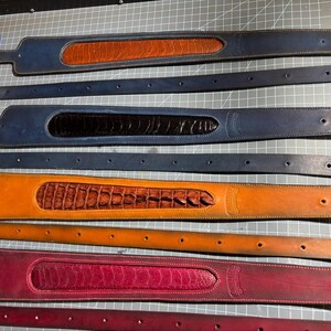 May include: Four leather belt sets arranged horizontally. Each set includes a wide, textured inlay strap and a matching plain strap with holes. Colors include blue with tan, blue with black, tan with brown, and magenta with dark red.
