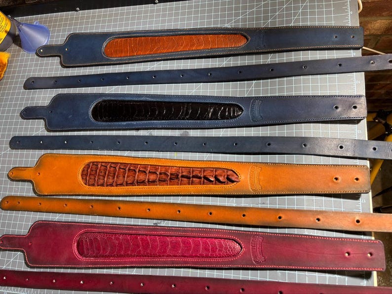 May include: Four leather belt sets arranged horizontally. Each set includes a wide, textured inlay strap and a matching plain strap with holes. Colors include blue with tan, blue with black, tan with brown, and magenta with dark red.