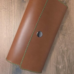 Puede incluir: Una cartera rectangular de cuero color canela con una solapa plegable asegurada con un bot&oacute;n de presi&oacute;n met&aacute;lico plateado. Los bordes de la cartera est&aacute;n acabados con costuras verdes visibles a lo largo de todo el per&iacute;metro.