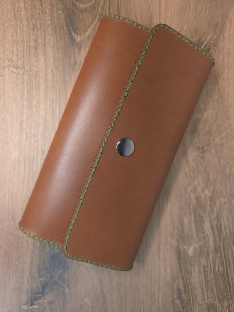 Puede incluir: Una cartera rectangular de cuero color canela con una solapa plegable asegurada con un bot&oacute;n de presi&oacute;n met&aacute;lico plateado. Los bordes de la cartera est&aacute;n acabados con costuras verdes visibles a lo largo de todo el per&iacute;metro.