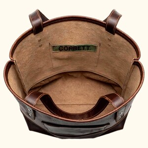 May include: A dark brown leather tote bag viewed from above, featuring two sturdy handles, a spacious interior with a tan suede-like lining, and a rectangular patch on the inner pocket displaying the text CORBETT in black lettering.