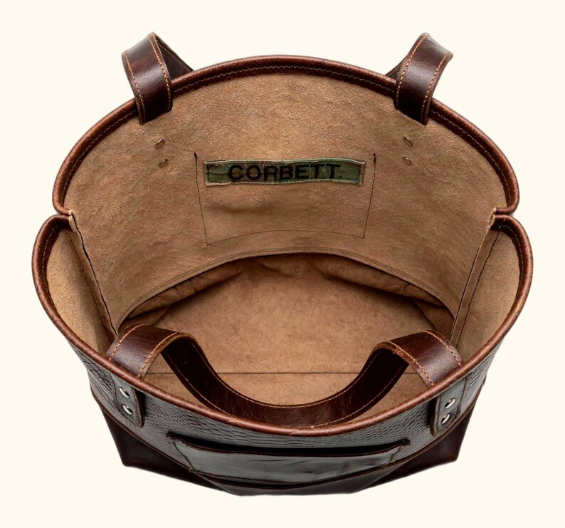 May include: A dark brown leather tote bag viewed from above, featuring two sturdy handles, a spacious interior with a tan suede-like lining, and a rectangular patch on the inner pocket displaying the text CORBETT in black lettering.