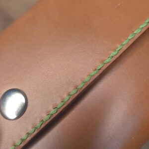 Puede incluir: Una cartera de cuero color canela con costuras de color verde brillante a lo largo del borde y un cierre de bot&oacute;n a presi&oacute;n de metal plateado. La superficie lisa del cuero contrasta con la apariencia texturizada y trenzada del hilo verde.