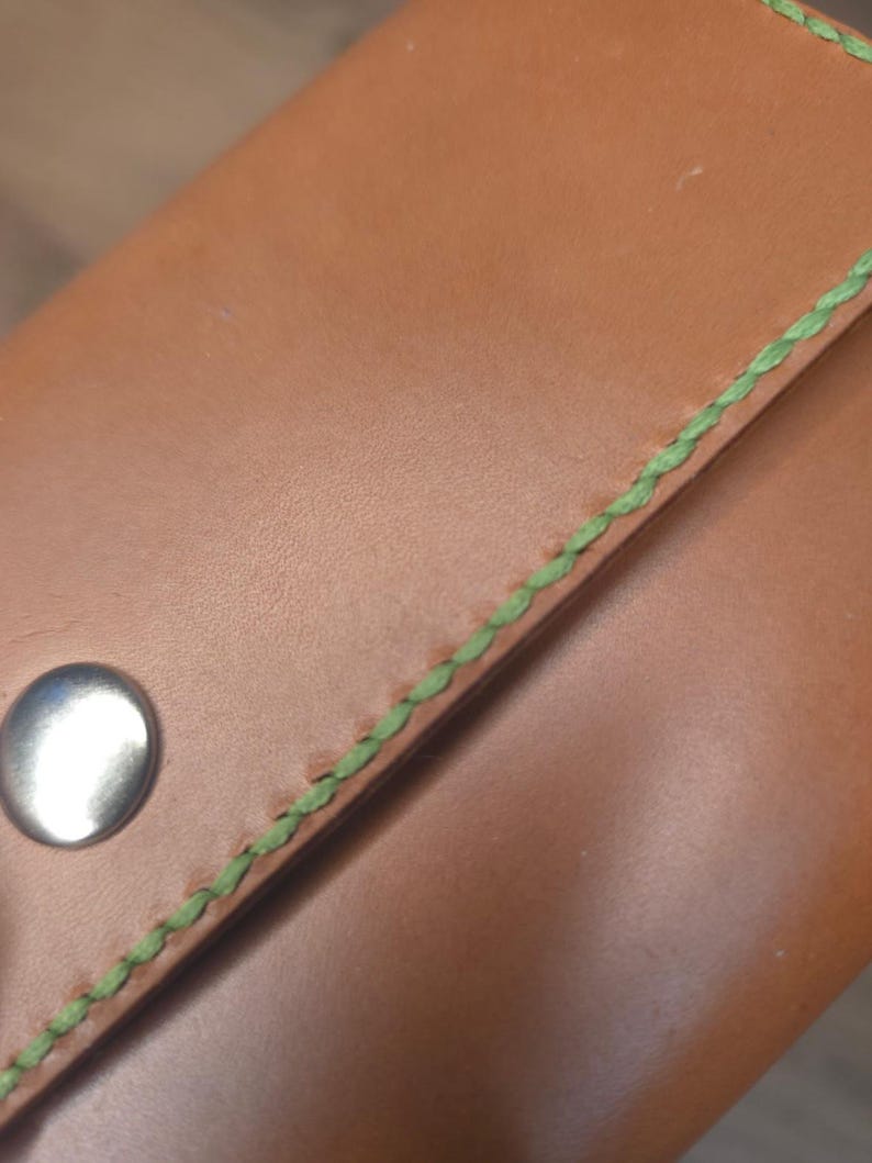 Puede incluir: Una cartera de cuero color canela con costuras de color verde brillante a lo largo del borde y un cierre de bot&oacute;n a presi&oacute;n de metal plateado. La superficie lisa del cuero contrasta con la apariencia texturizada y trenzada del hilo verde.