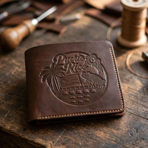 Puede incluir: Una cartera de cuero marrón oscuro con un diseño circular grabado en la parte frontal. El diseño incluye el texto Puerto Rico, una palmera, una bandera, una rana coquí y una flor de hibisco, cosido con hilo de color marrón claro.