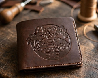 May include: A dark brown leather bifold wallet featuring an embossed circular design on the front. The design includes the text Puerto Rico, a palm tree, a flag, a coqui frog, and a hibiscus flower, all stitched with light brown thread.