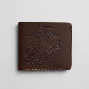 May include: A dark brown leather bifold wallet featuring an embossed United States Marine Corps Eagle, Globe, and Anchor emblem on the front cover. The wallet is constructed with visible brown stitching along the edges and rounded corners.