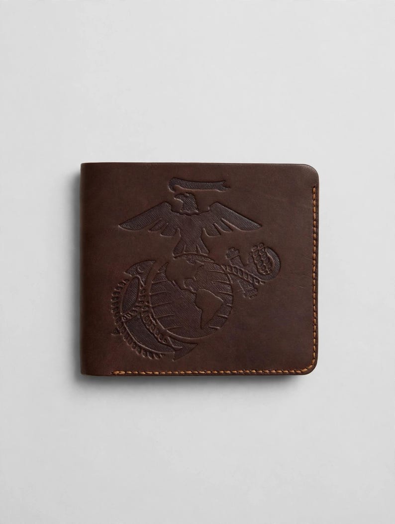 May include: A dark brown leather bifold wallet featuring an embossed United States Marine Corps Eagle, Globe, and Anchor emblem on the front cover. The wallet is constructed with visible brown stitching along the edges and rounded corners.