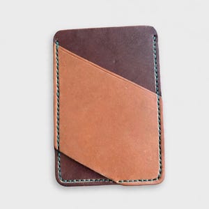 May include: A rectangular leather card holder featuring a dark brown base layer and a lighter tan diagonal pocket overlay. The edges are secured with visible teal stitching, creating a contrast against the two-toned brown leather surface.