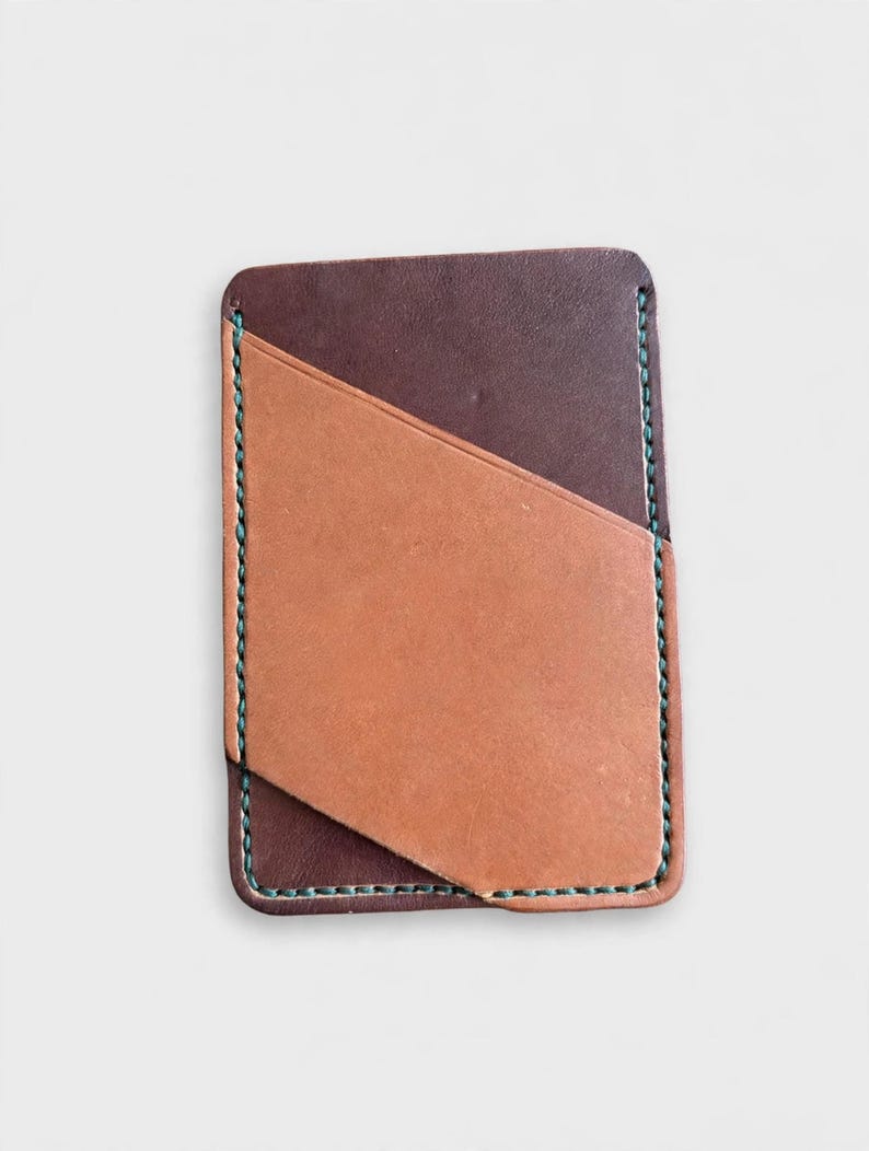 May include: A rectangular leather card holder featuring a dark brown base layer and a lighter tan diagonal pocket overlay. The edges are secured with visible teal stitching, creating a contrast against the two-toned brown leather surface.