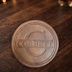 May include: A round, tan leather coaster featuring an embossed design with a large capital letter C in the background and the name CORBETT printed horizontally across the center within a double-lined circular border.