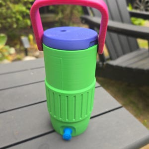 May include: A colorful water cooler with a pink handle, a blue lid, and a green body. The cooler has a blue spigot at the bottom. The design is modern and functional, perfect for outdoor use or picnics.