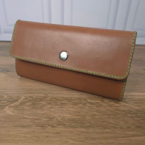 Puede incluir: Una cartera rectangular de cuero color canela con un cierre de bot&oacute;n a presi&oacute;n de metal plateado. Los bordes de la cartera est&aacute;n cosidos con hilo grueso de color verde lima brillante, creando un contraste marcado con la superficie de cuero liso.