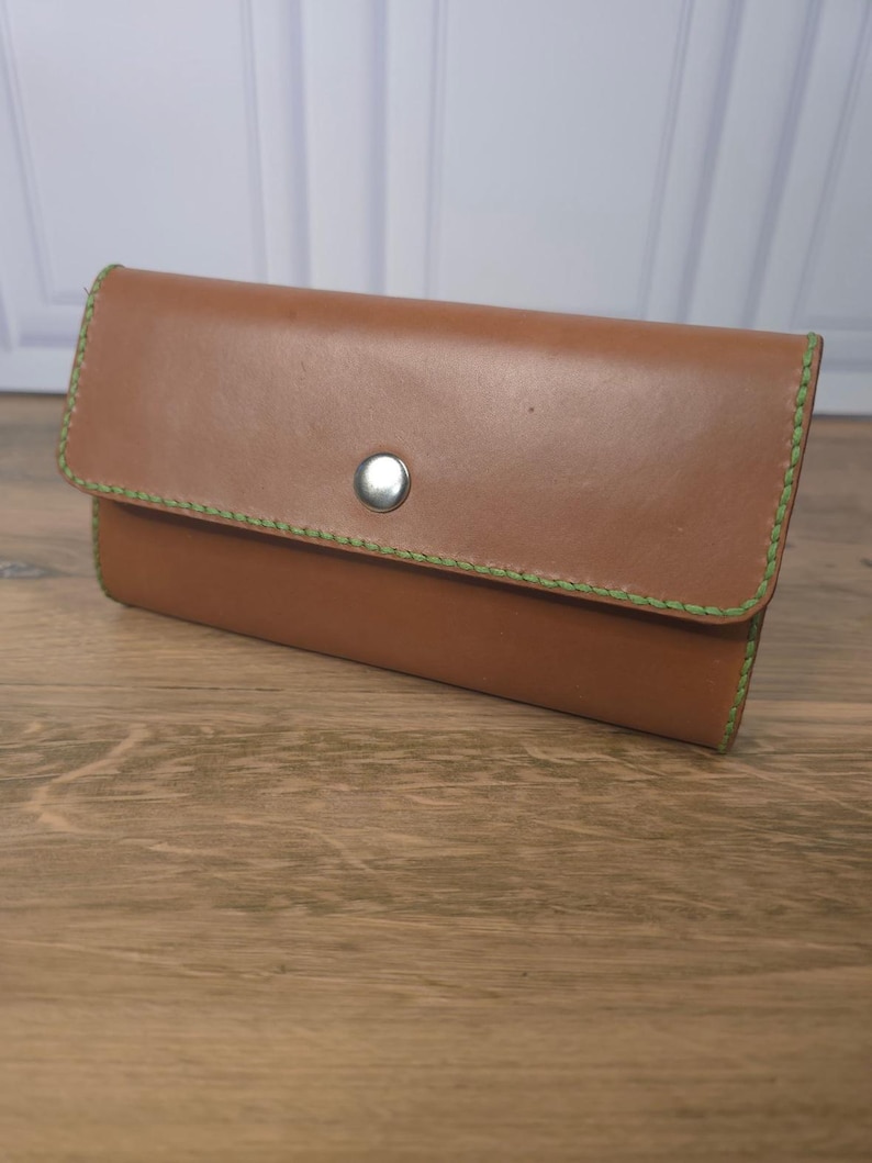 Puede incluir: Una cartera rectangular de cuero color canela con un cierre de bot&oacute;n a presi&oacute;n de metal plateado. Los bordes de la cartera est&aacute;n cosidos con hilo grueso de color verde lima brillante, creando un contraste marcado con la superficie de cuero liso.