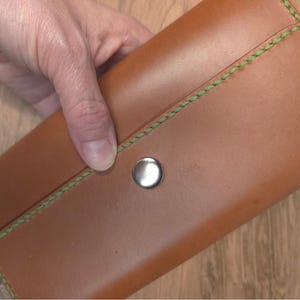 Puede incluir: Una cartera rectangular de cuero color canela con una superficie lisa y un cierre de bot&oacute;n a presi&oacute;n de metal plateado. Los bordes de la cartera est&aacute;n asegurados con costuras verdes gruesas y visibles que bordean la solapa.