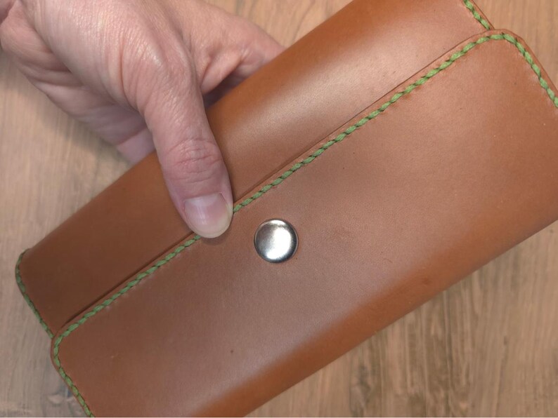 Puede incluir: Una cartera rectangular de cuero color canela con una superficie lisa y un cierre de bot&oacute;n a presi&oacute;n de metal plateado. Los bordes de la cartera est&aacute;n asegurados con costuras verdes gruesas y visibles que bordean la solapa.