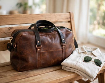 May include: A brown leather duffel bag with black leather handles and trim, featuring copper-toned metal hardware. Beside the bag lies a folded light-colored button-down shirt and a pair of gold-framed aviator sunglasses with dark lenses.
