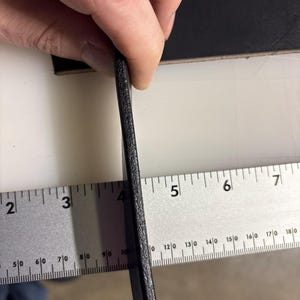 May include: A thick, black leather strip held vertically against a metal ruler. The ruler shows the leather edge measures approximately 0.25 inches in thickness. The leather has a textured, matte surface and a solid, dark black color throughout.