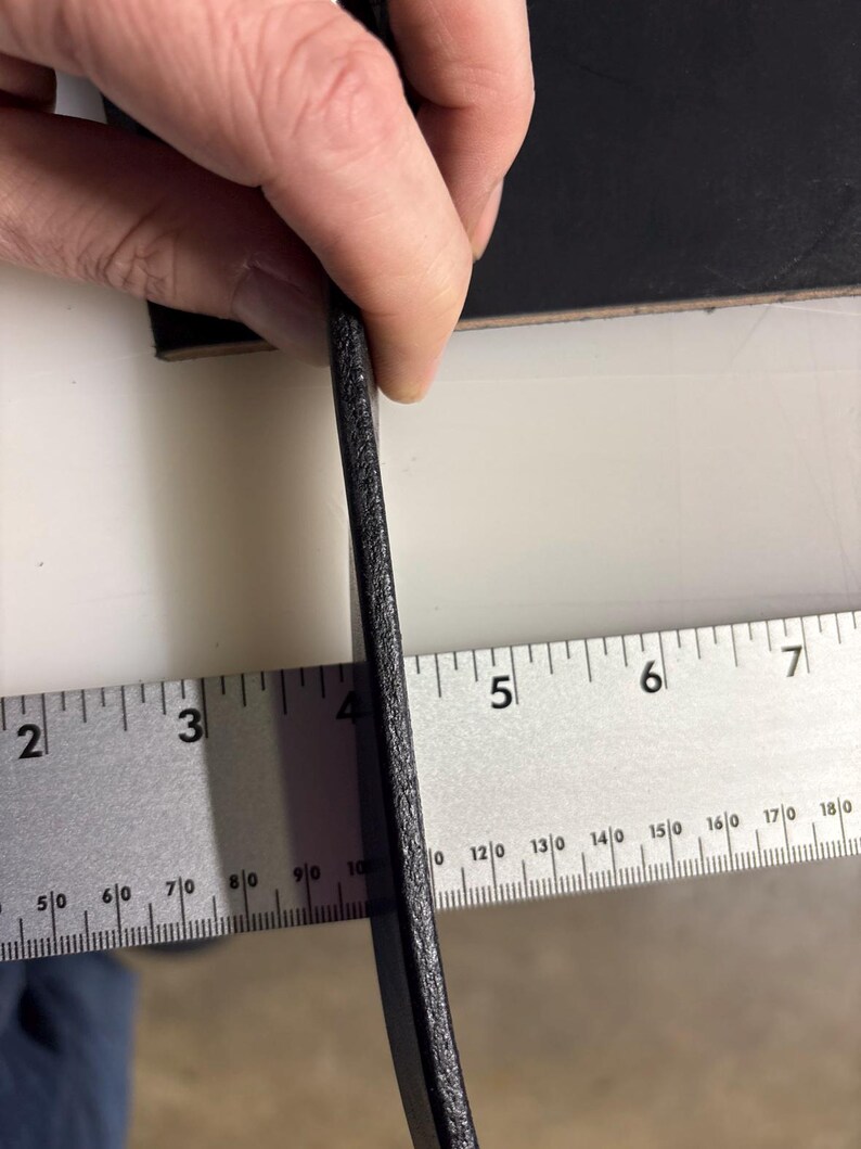 May include: A thick, black leather strip held vertically against a metal ruler. The ruler shows the leather edge measures approximately 0.25 inches in thickness. The leather has a textured, matte surface and a solid, dark black color throughout.