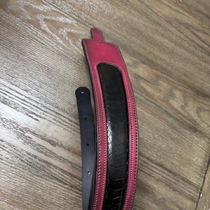May include: A guitar strap featuring a wide, padded section with a black, textured, reptilian-patterned center panel bordered by vibrant pink leather edges. The strap includes a narrow, smooth black leather adjustment tail with punched holes.