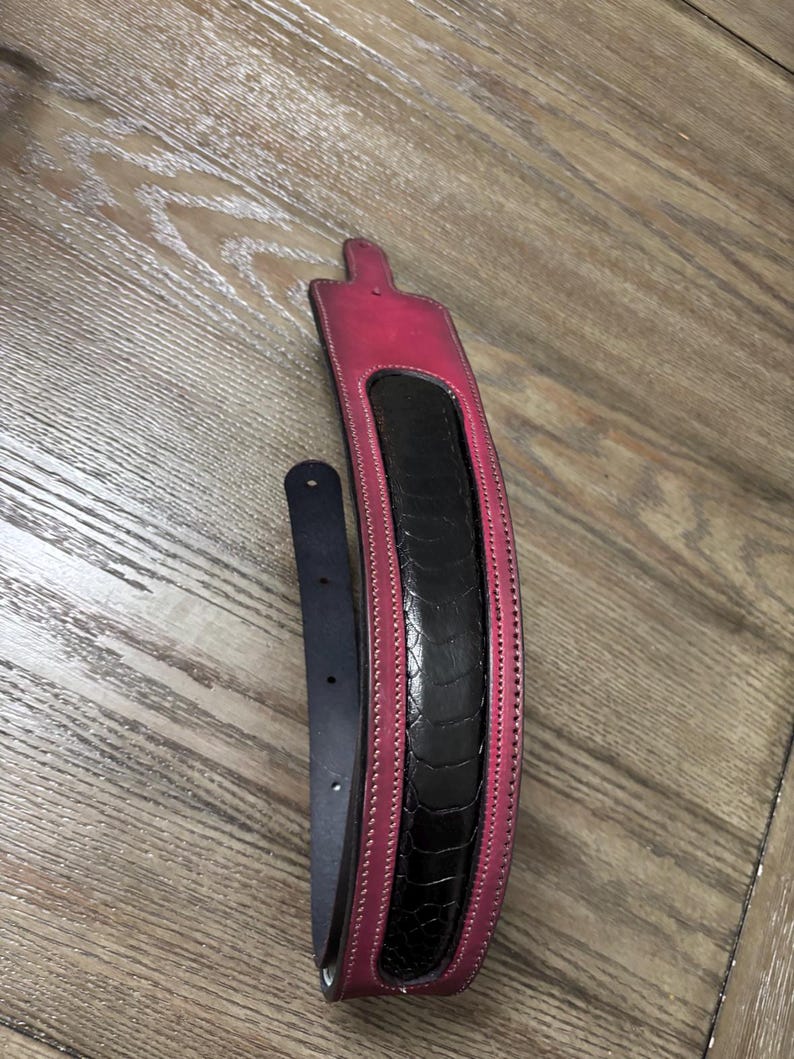 May include: A guitar strap featuring a wide, padded section with a black, textured, reptilian-patterned center panel bordered by vibrant pink leather edges. The strap includes a narrow, smooth black leather adjustment tail with punched holes.