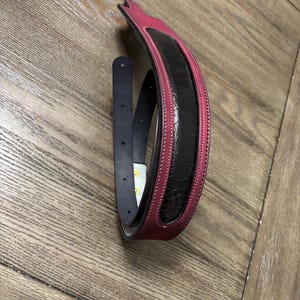 May include: A belt featuring a deep magenta leather outer band with white stitching and a central black textured panel with a reptilian-like scale pattern. The belt includes a dark blue inner strap with visible adjustment holes and gold-toned hardware.