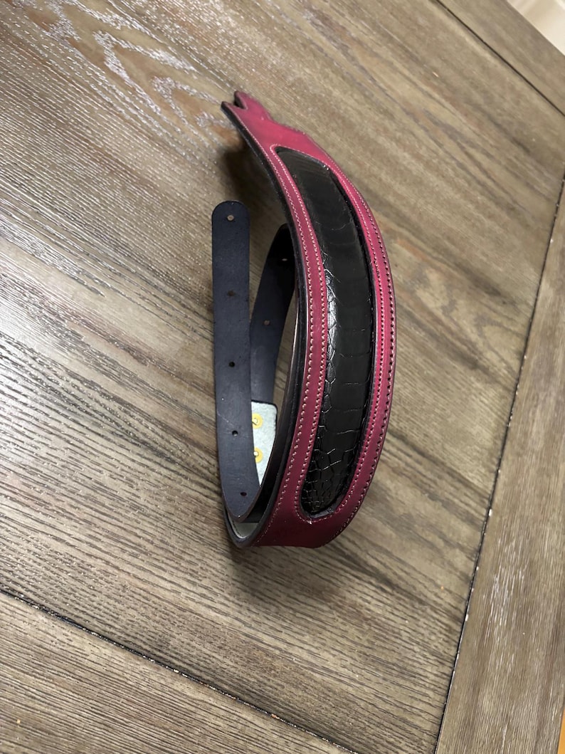May include: A belt featuring a deep magenta leather outer band with white stitching and a central black textured panel with a reptilian-like scale pattern. The belt includes a dark blue inner strap with visible adjustment holes and gold-toned hardware.